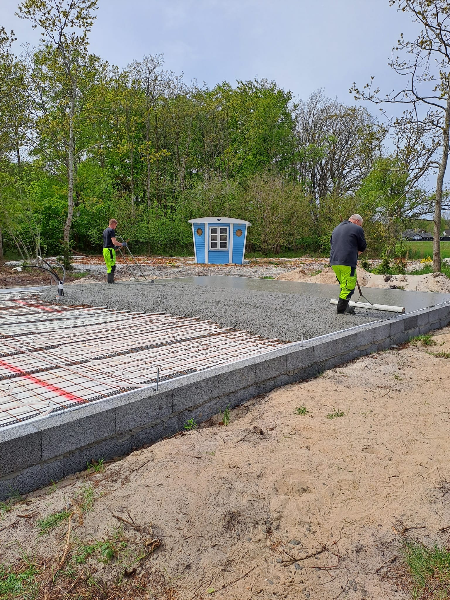 Arbejder udjævner betonfundament ved lille blåt skur
