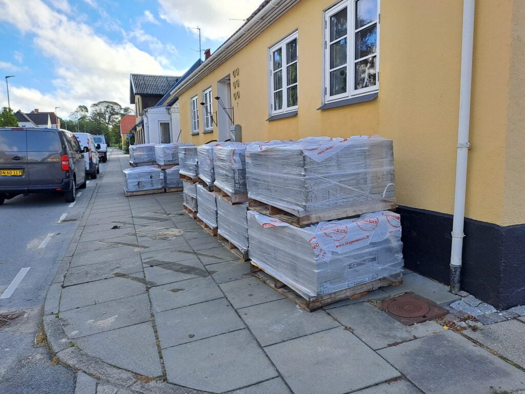 Paller med byggematerialer foran gult hus på fortov